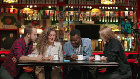 Portrait-of-cheerful-young-friends-looking-at-smart-phone-while-sitting-in-cafe.-Mixed-race-people-sitting-at-a-table-in-restaurant-using-mobile-phone.